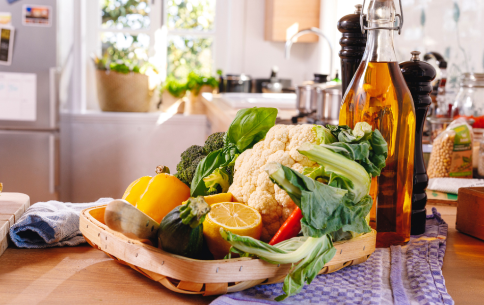 Blumenkohl, Zitrone und Zucchini liegen in einem geflochtenen Korb in einer Küche, hinter dem Korb steht eine Flasche Öl, Salz- und Pfeffermühlen
